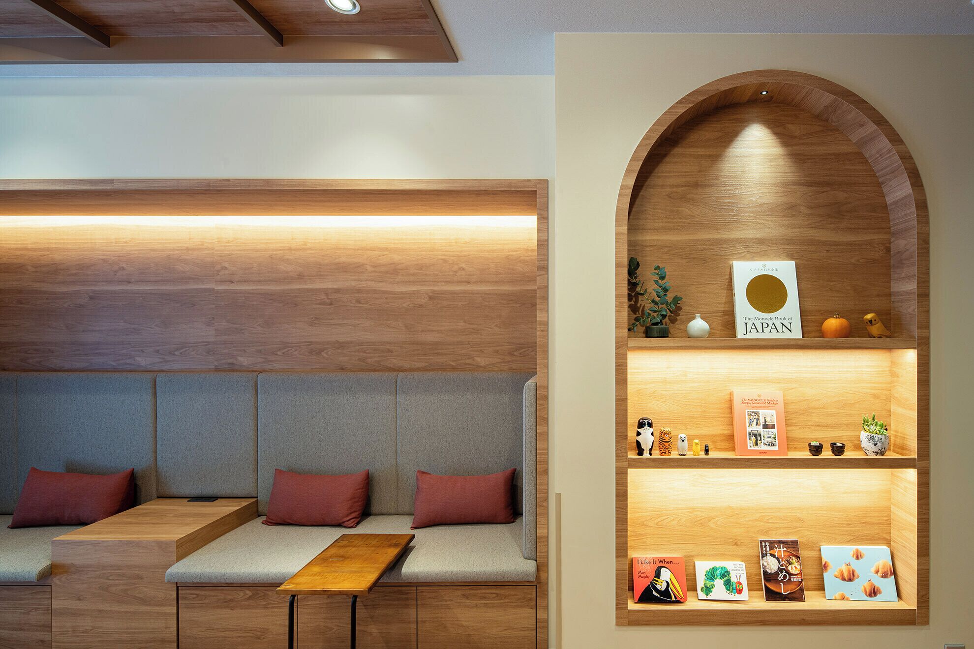 A modern seating area with a bench and cushions, a wooden bookshelf with books and decorative items, and a wooden ceiling with recessed lighting.