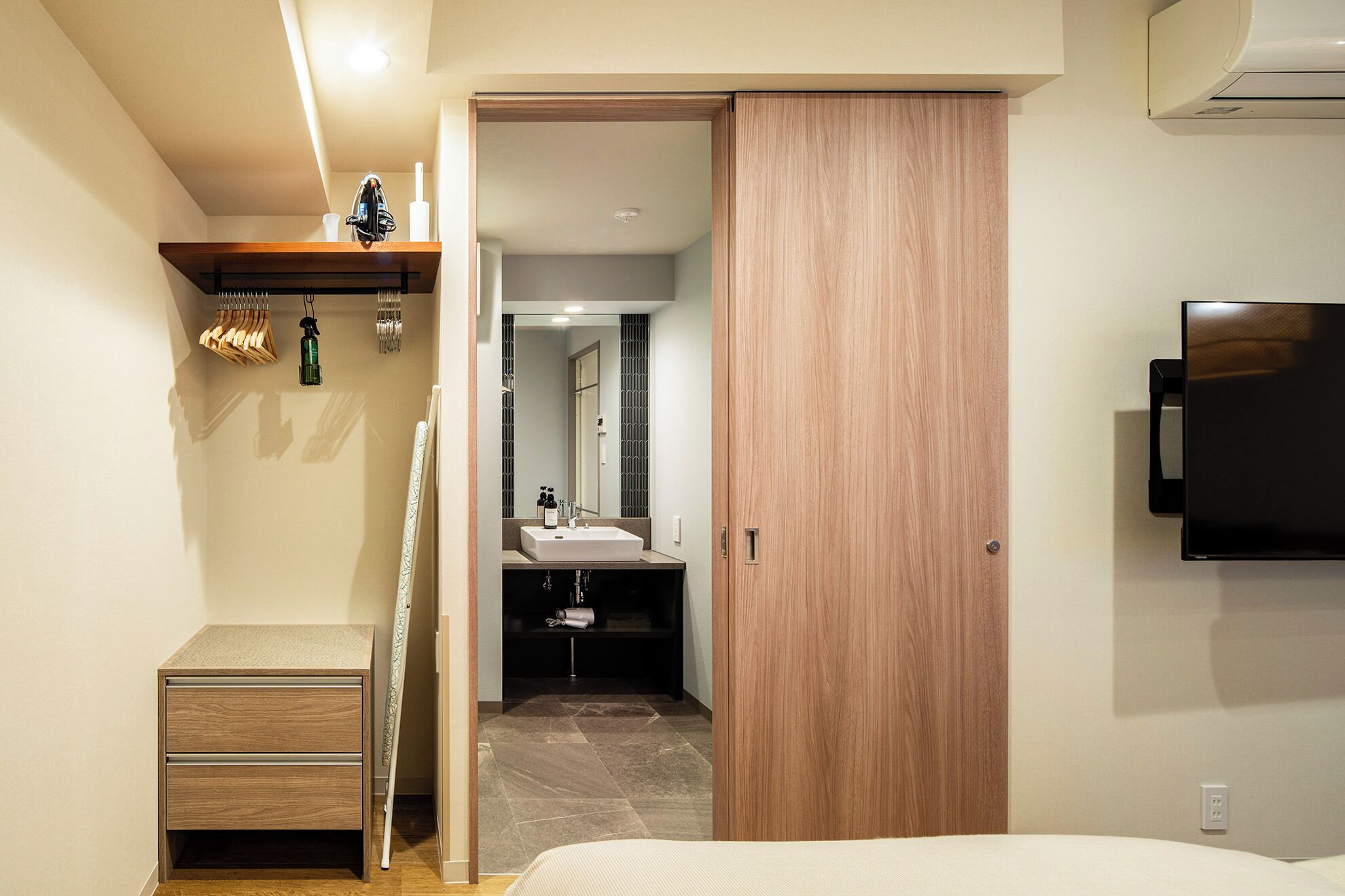 A modern bedroom with a wooden wardrobe, a bedside table, a TV mounted on the wall, and a bathroom visible through an open door.