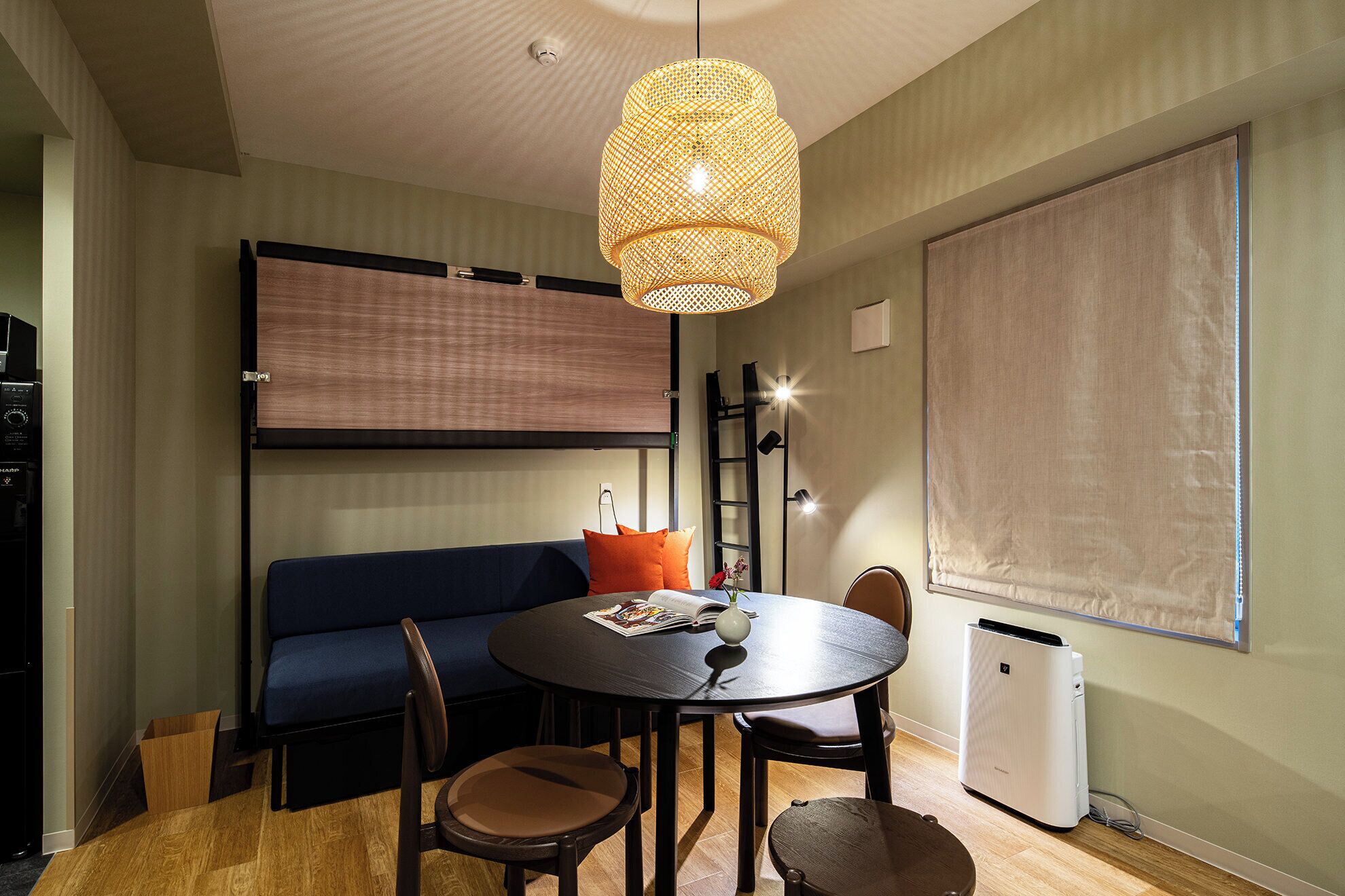 A compact living space with a sofa, a round table, and chairs. A large window with a blind is on the right, and a ceiling-mounted light fixture is above the table.