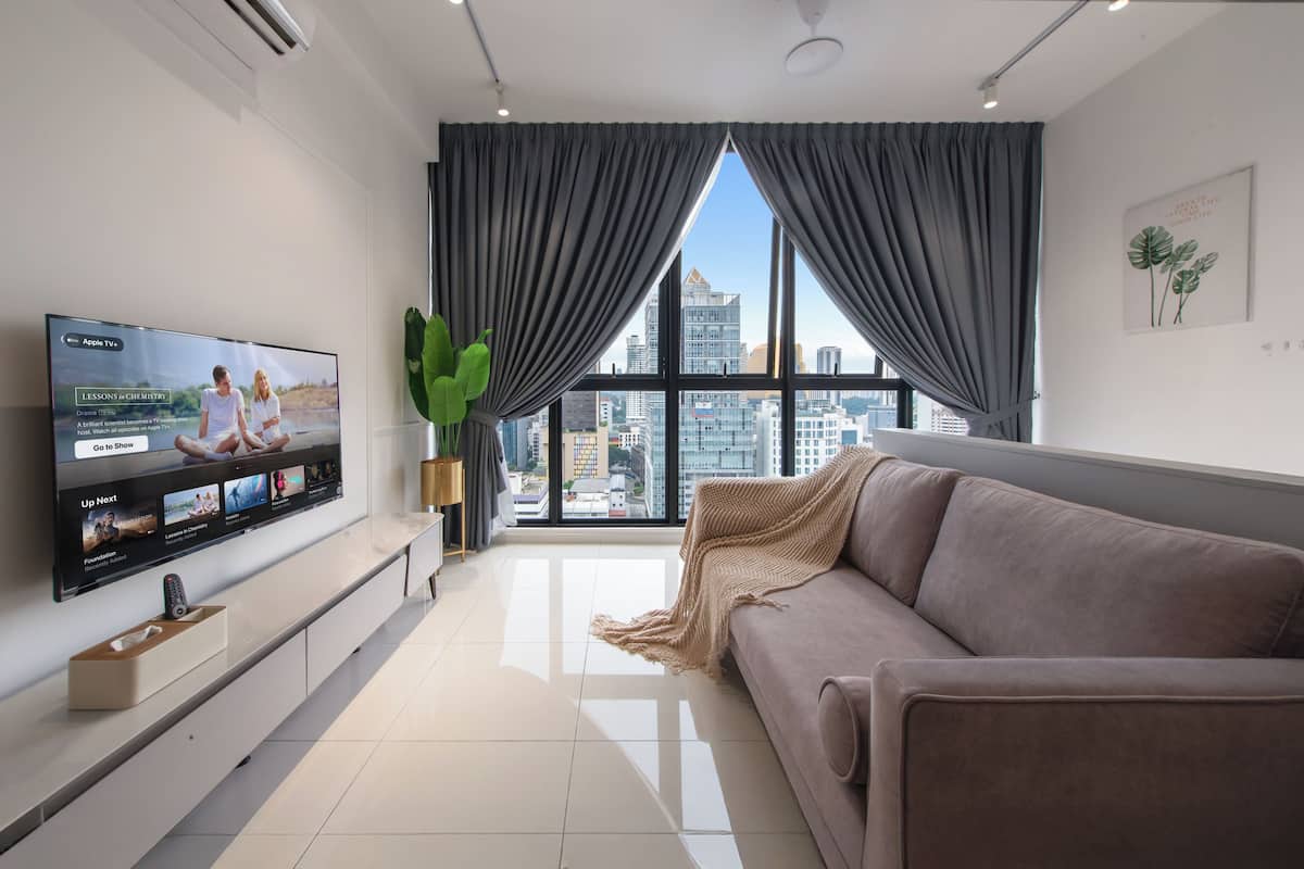 A modern living room with a large flat-screen TV, a sofa, a plant, and a city view through large windows.