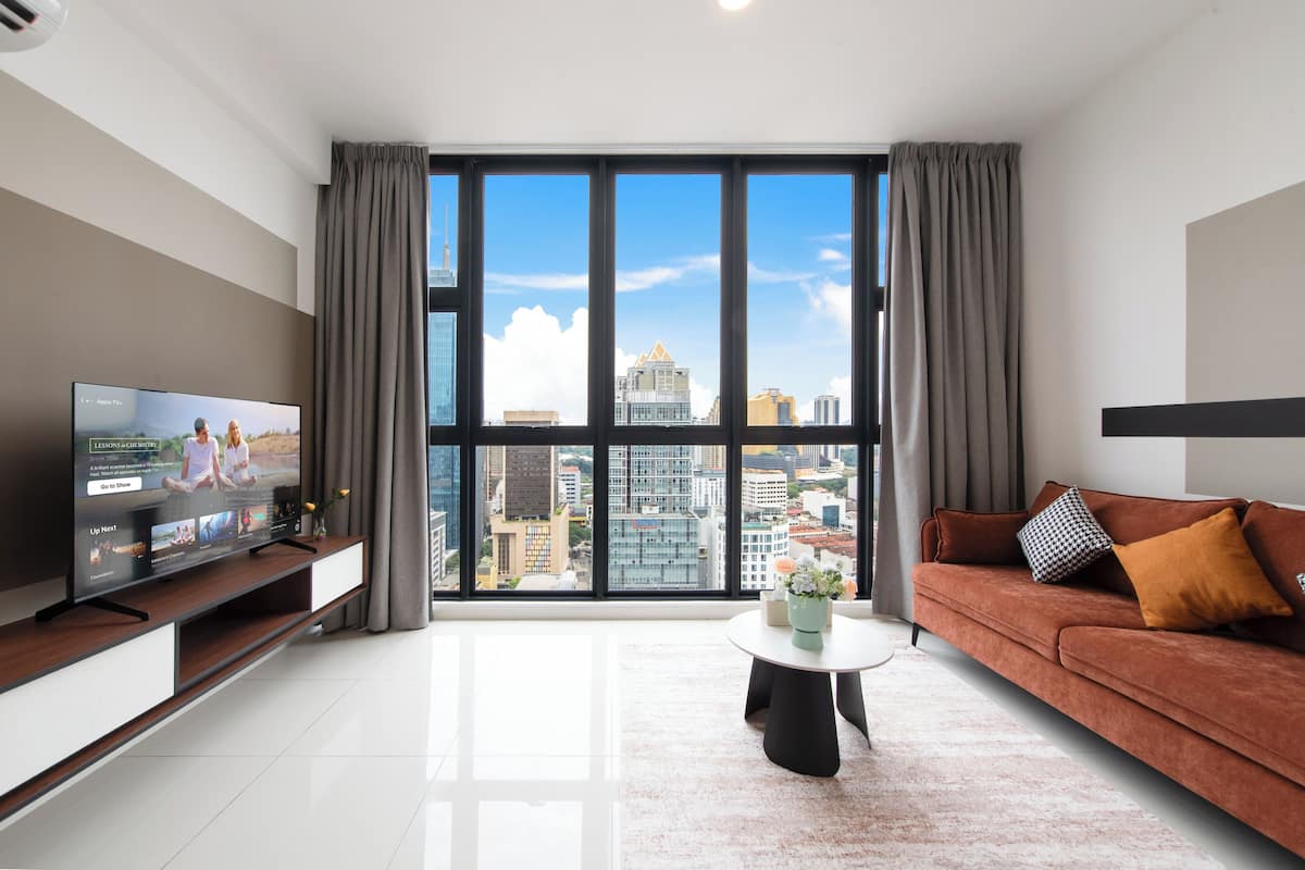 A modern living room with a large TV on a wooden stand, a brown sofa with cushions, a round coffee table, and a view of city buildings through large windows.