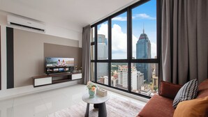 A modern living room with a large window, a flat-screen TV, a small round table with a vase, and a sofa with cushions. - Majestic by Avery &10 mins to Twin Tower (Kuala Lumpur)