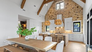 A modern kitchen with a stone wall, wooden beams, white cabinets, and a dining area with a wooden table and benches. - Luxe Lake Martin Home  Pool Hot Tub  Boat Dock (Dadeville)