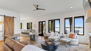 A spacious living area with a large wooden table, a round coffee table, and a sofa. There are multiple windows offering views of a body of water. - Luxe Lake Martin Home  Pool Hot Tub  Boat Dock (Dadeville)