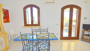 A dining area with a table and chairs, two arched windows, an air conditioning unit, and a television on a stand.