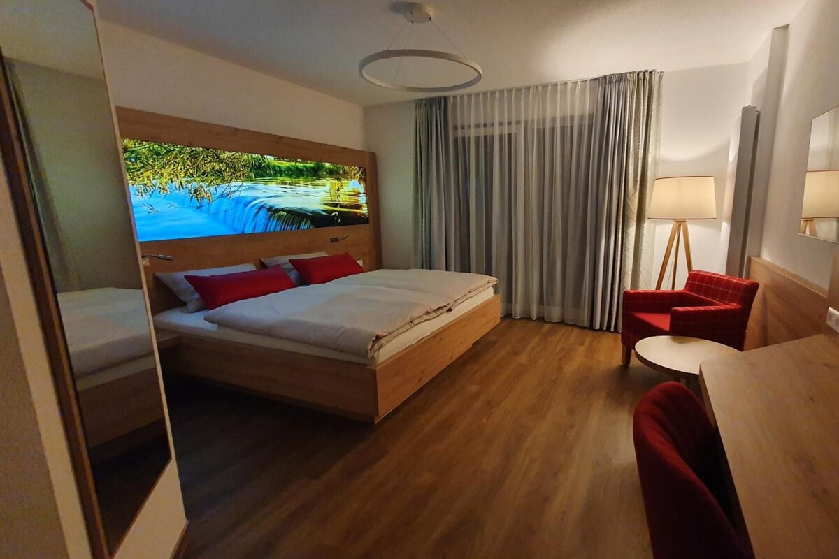 A hotel room with a bed, a desk, a chair, a lamp, a mirror, and a view of a natural landscape.