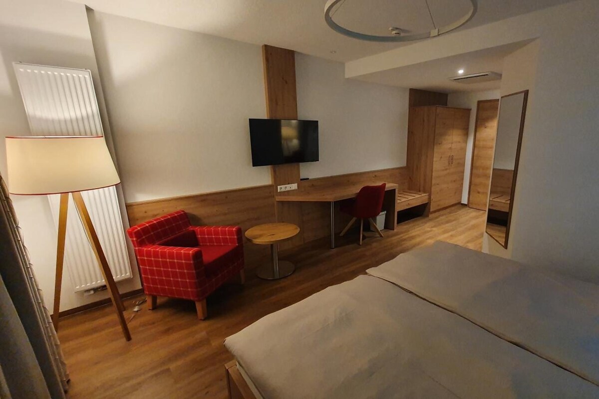 A hotel room with a bed, a TV, a red chair, a small table, and a lamp.
