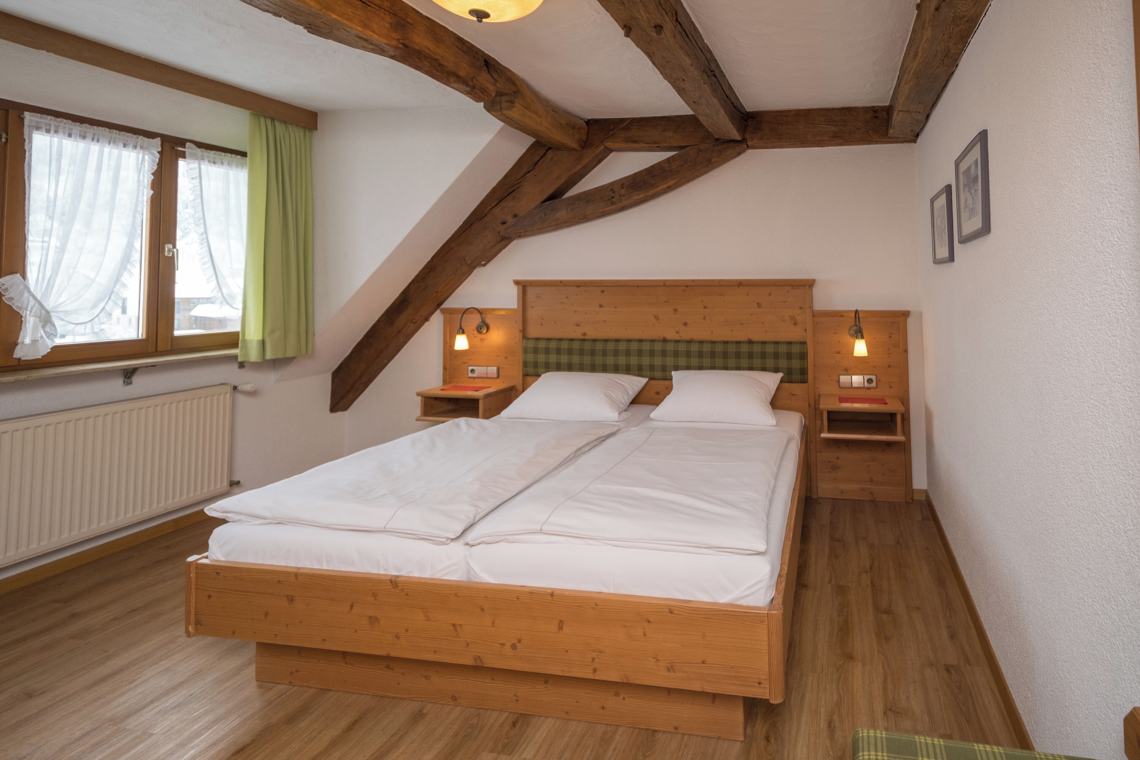 A bedroom with a wooden bed, two bedside tables, a window with curtains, and a radiator.