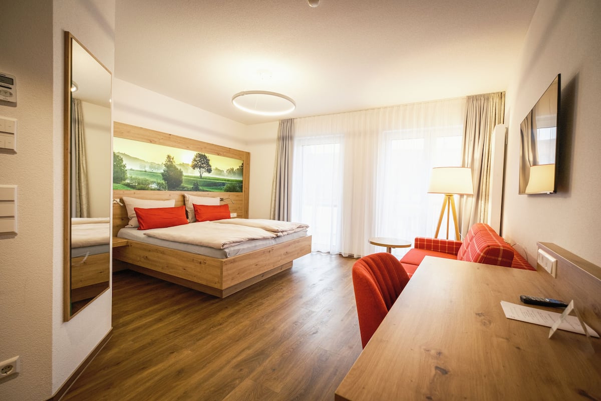 Ein Hotelzimmer mit einem hölzernen Bett, einem Holztisch, einem roten Sessel, einer Lampe und einem Gemälde an der Wand.