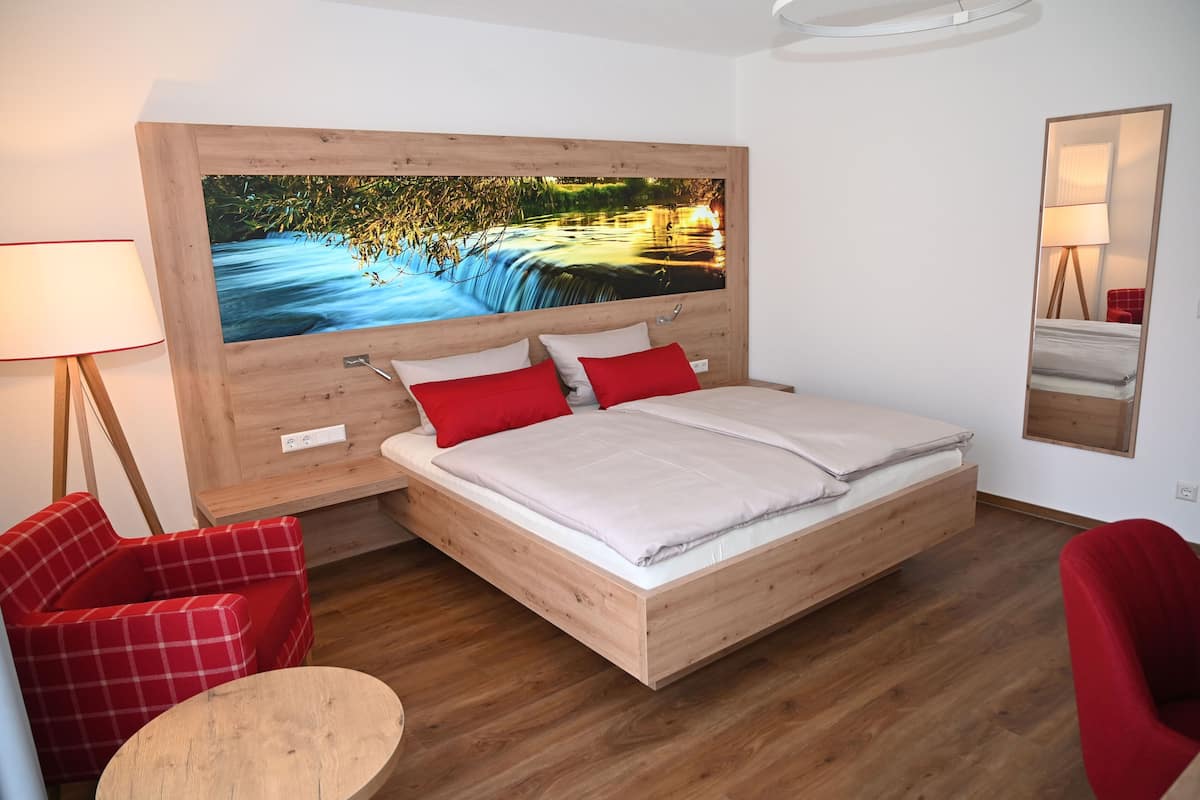 Ein modernes Schlafzimmer mit einem hölzernen Kopfteil, einem großen Bett, roten und weißen Kissen, einem roten Sessel, einem kleinen runden Tisch und einem großen Wandbild mit einer Flusslandschaft.