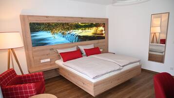 A modern bedroom with a wooden headboard, a large bed, red and white pillows, a red chair, a small round table, and a large wall-mounted painting of a river scene.