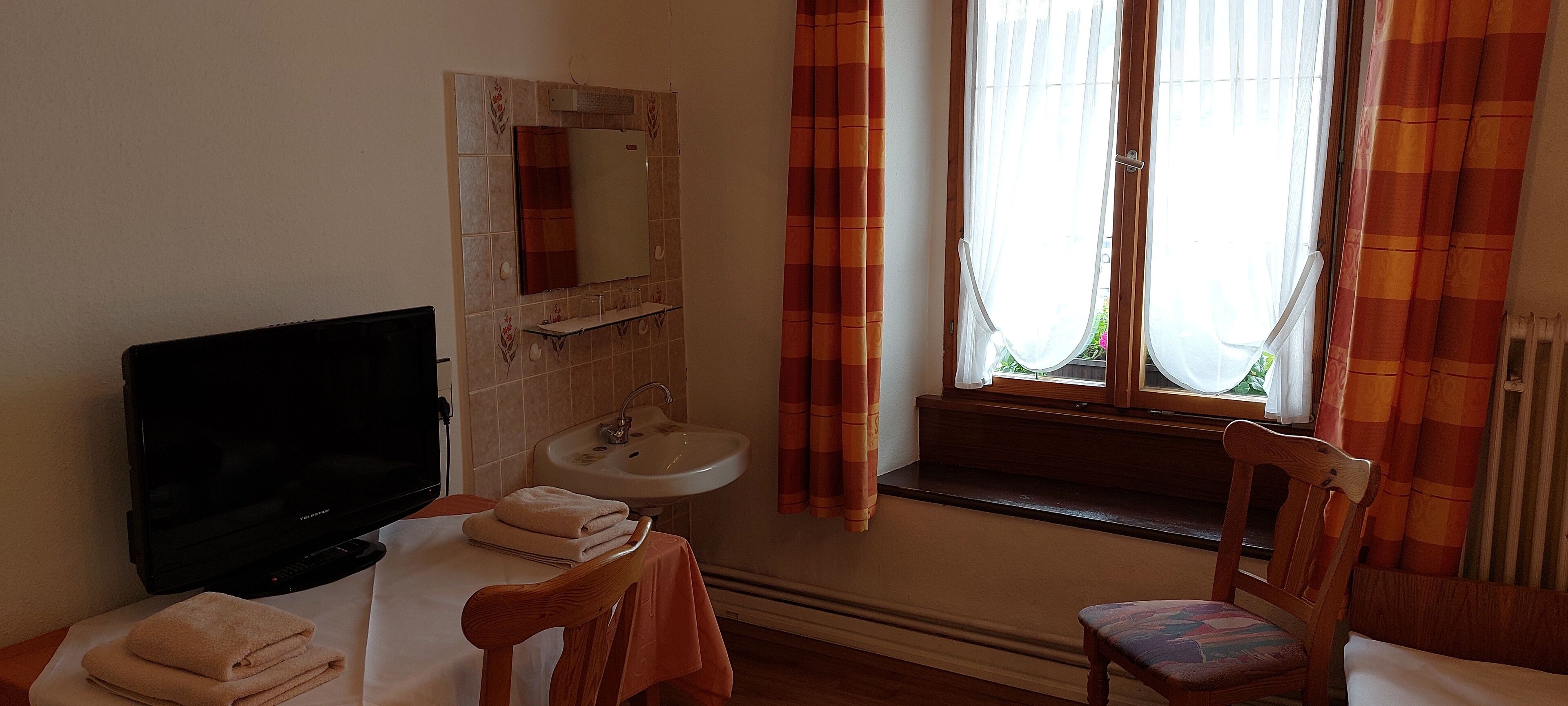 Una habitación con televisión, lavamanos, espejo, ventana con cortinas y una silla.