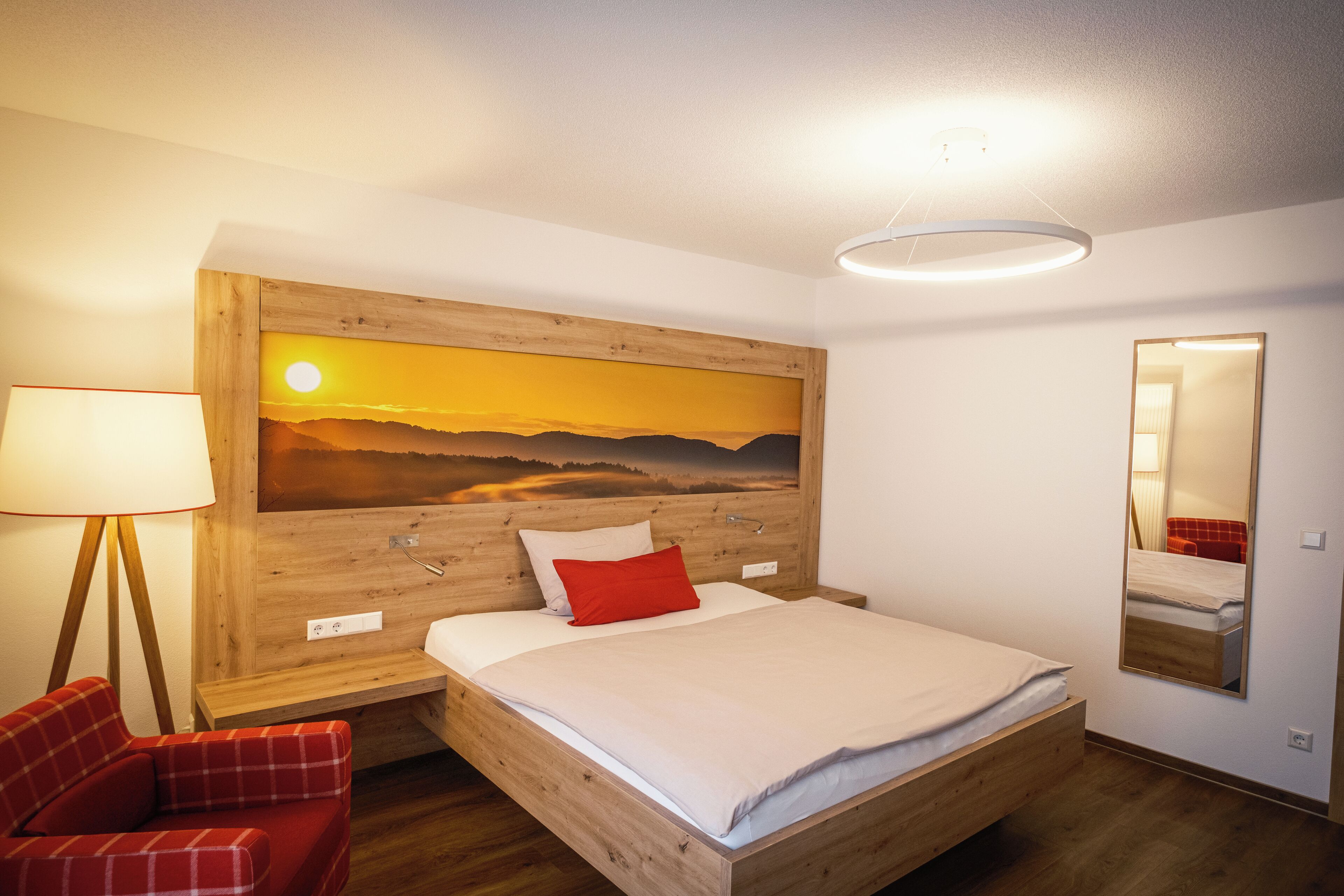 Un dormitorio con cabecera de madera, cama con ropa de cama blanca y roja, un sillón rojo a cuadros, una mesita de noche y un espejo montado en la pared.