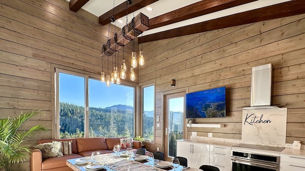 A modern kitchen with a wooden interior, a dining area with a long table set for six, and a large window offering a scenic mountain view. - Sunset Lodge - Modern Mountain Luxury - Epic Views - Sleeps 6 (Comox Valley C (Puntledge-B*)