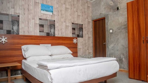 A bedroom with a wooden bed, a small bedside table, a wall-mounted fan, and a wooden wardrobe. - Avanti retreat  (Vaadhoo)