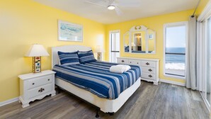 A bedroom with a bed, bedside tables, a ceiling fan, a window with a view of the ocean, and a painting on the wall.