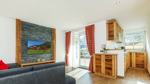 A modern living room with a grey sofa, a flat-screen TV mounted on a textured wall, and a wooden cabinet in the kitchen area. - Reifelberg 45 sqm, terrace, kitchen, 1 bedroom, living room, max 3 pers, WLAN (Ruhpolding)