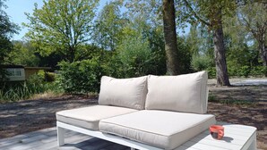 Ein weißes Outdoor-Sofa mit Kissen auf einer gepflasterten Terrasse, ein kleiner Tisch mit einer Tasse, umgeben von Grünpflanzen und Bäumen.