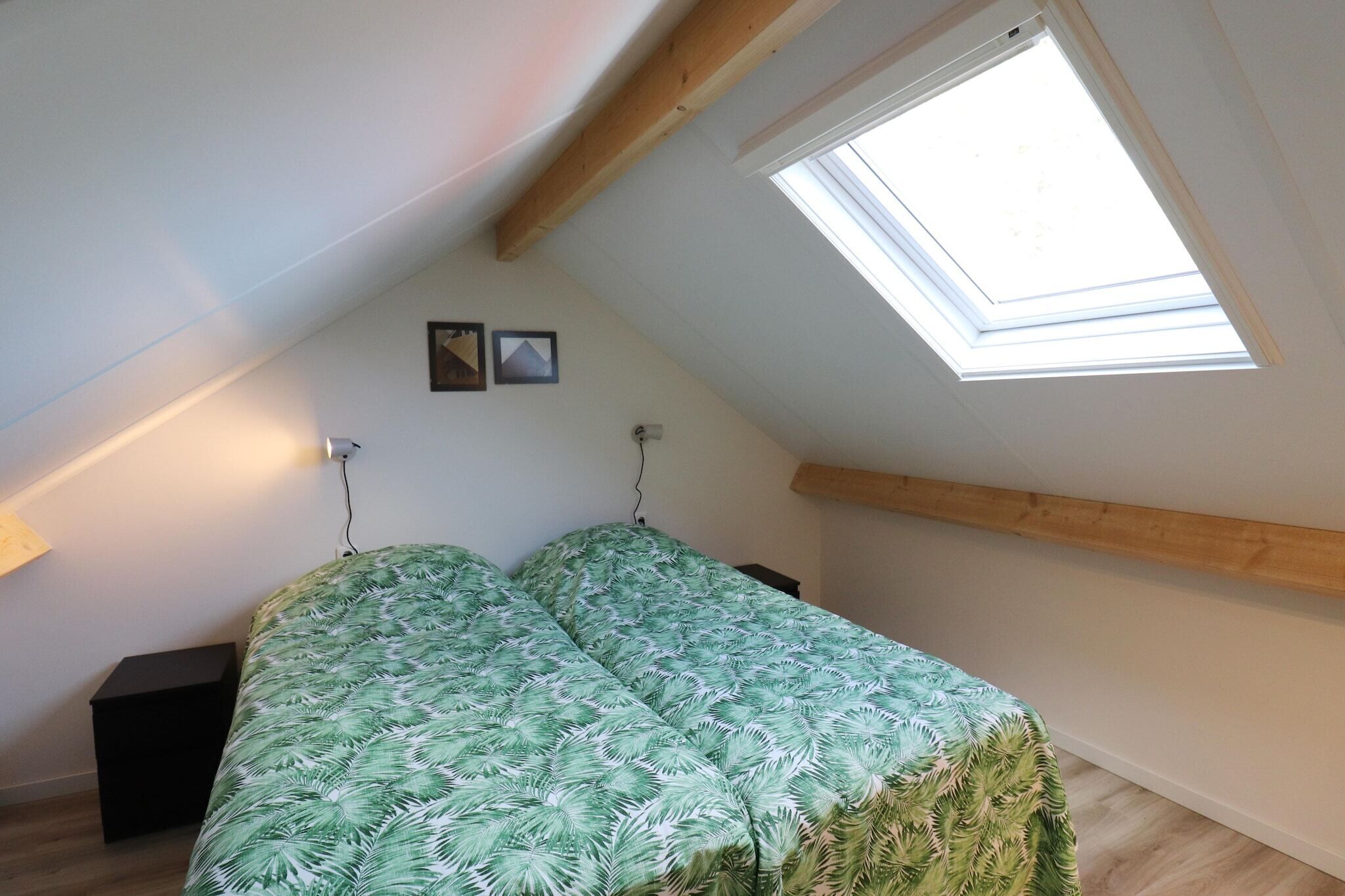 A bedroom with a sloped ceiling, a skylight, a bed with a green leaf-patterned bedspread, a bedside table, and a wall-mounted lamp.