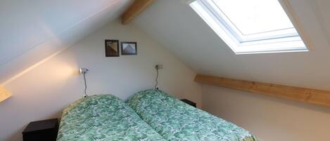A bedroom with a sloped ceiling, a skylight, a bed with a green leaf-patterned bedspread, a bedside table, and a wall-mounted lamp.