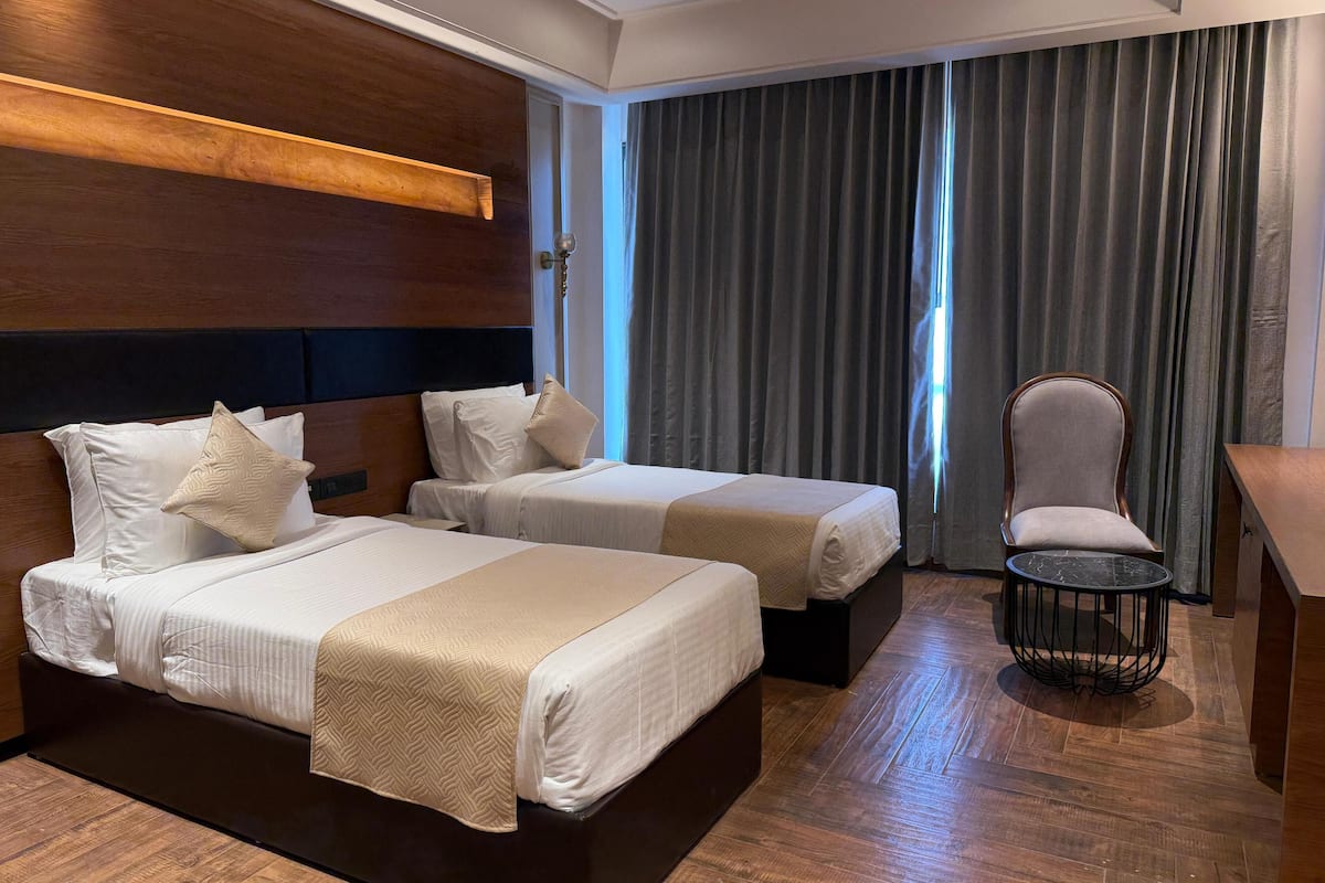 Quarto de hotel com duas camas, uma cadeira, uma mesinha e vista para o exterior através das cortinas.