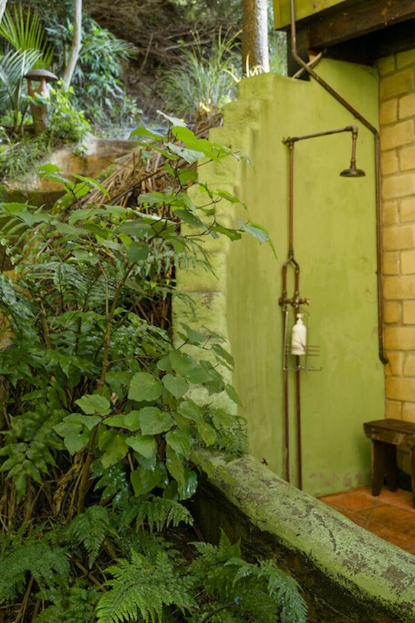 A rustic outdoor shower area with a green wall, a wooden bench, and lush greenery.