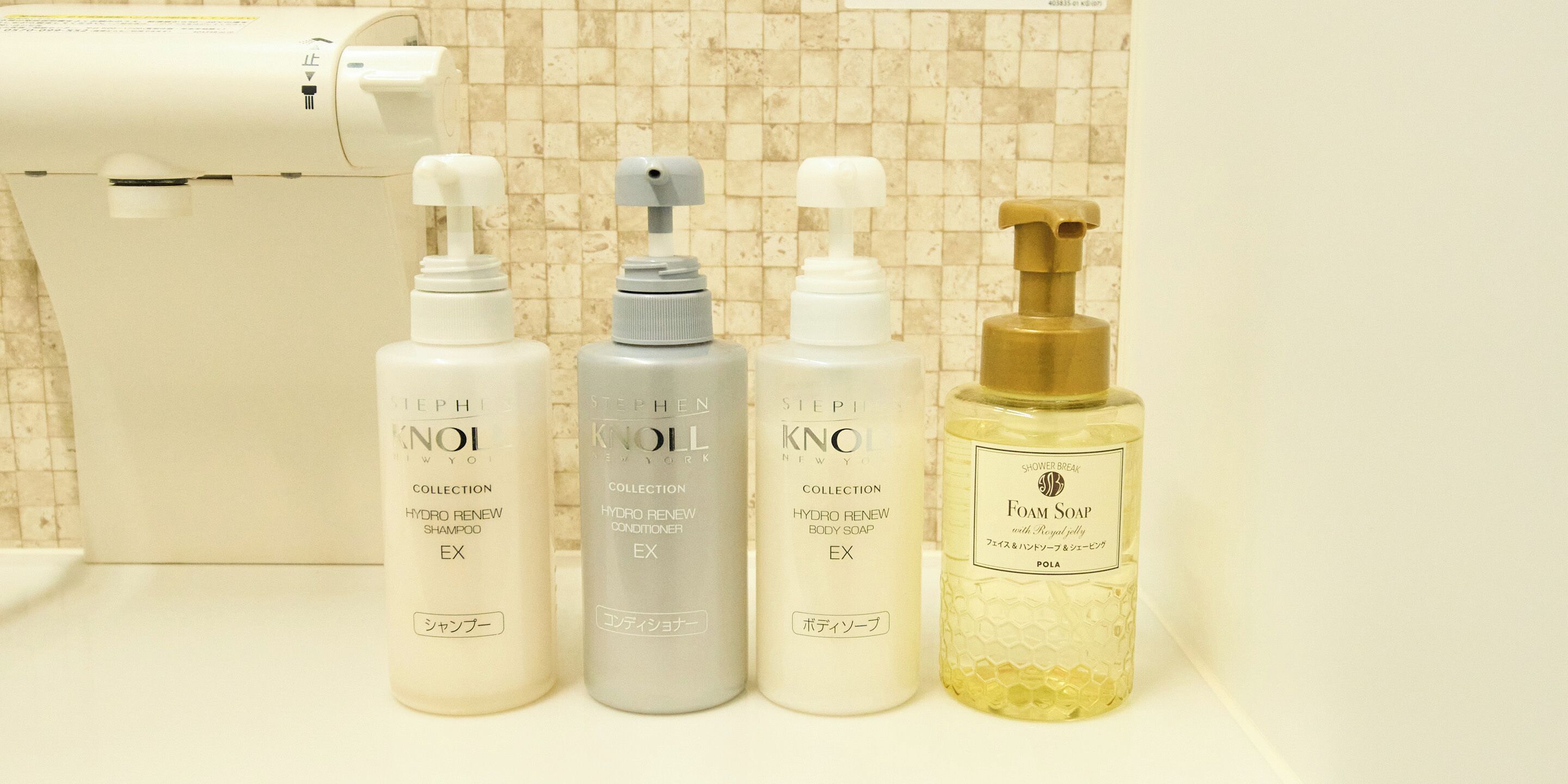 Une salle de bain équipée d’un sèche-mains, de quatre flacons de produits de toilette et d’un mur recouvert de carreaux.