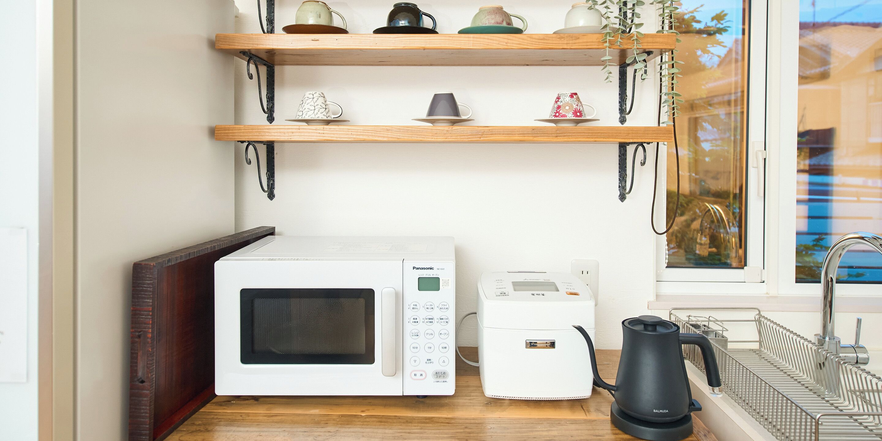 Una zona de cocina con microondas, cocedor de arroz y tetera sobre la encimera. Hay estantes de madera arriba con diversos utensilios de cocina.