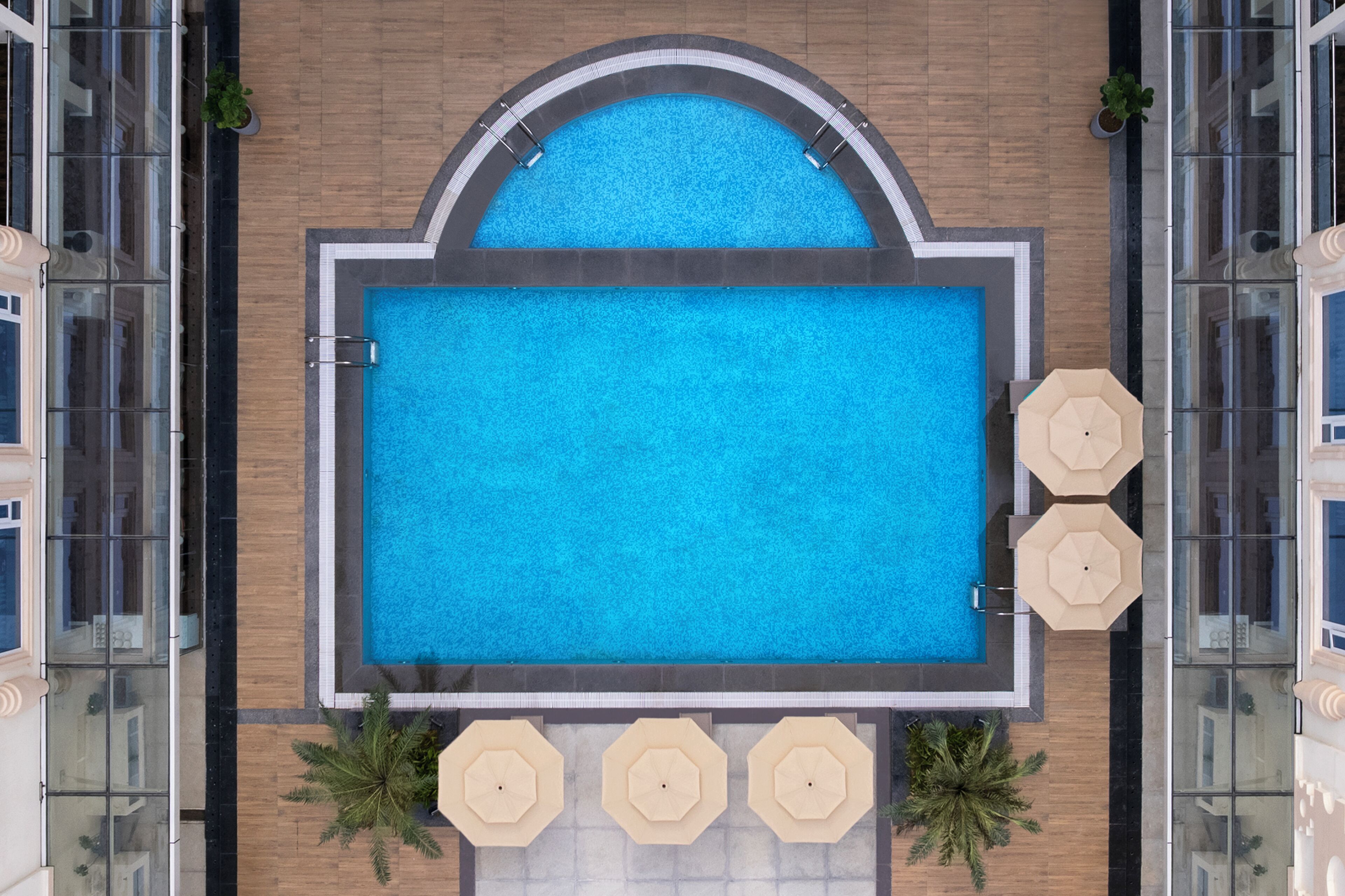Vista aérea de una piscina rectangular con solárium empedrado, rodeada por tres sombrillas beige y plantas en macetas.