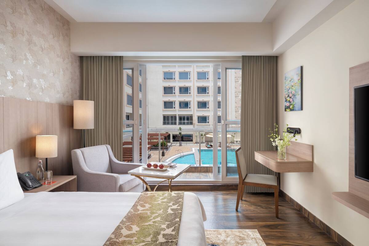 Une chambre d’hôtel avec un lit, un bureau, une chaise, une télévision et une fenêtre donnant sur une piscine et un immeuble.