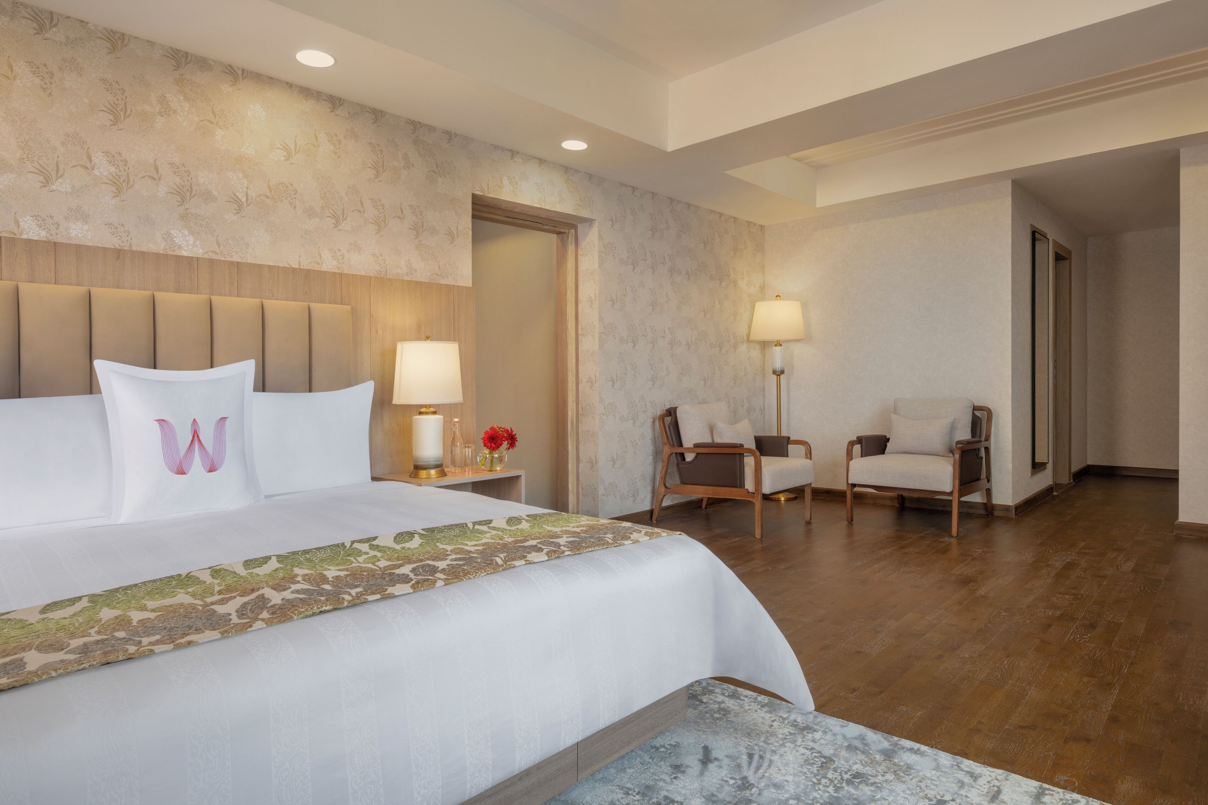 A hotel room with a large bed, two chairs, a nightstand with a lamp, and a wooden headboard.