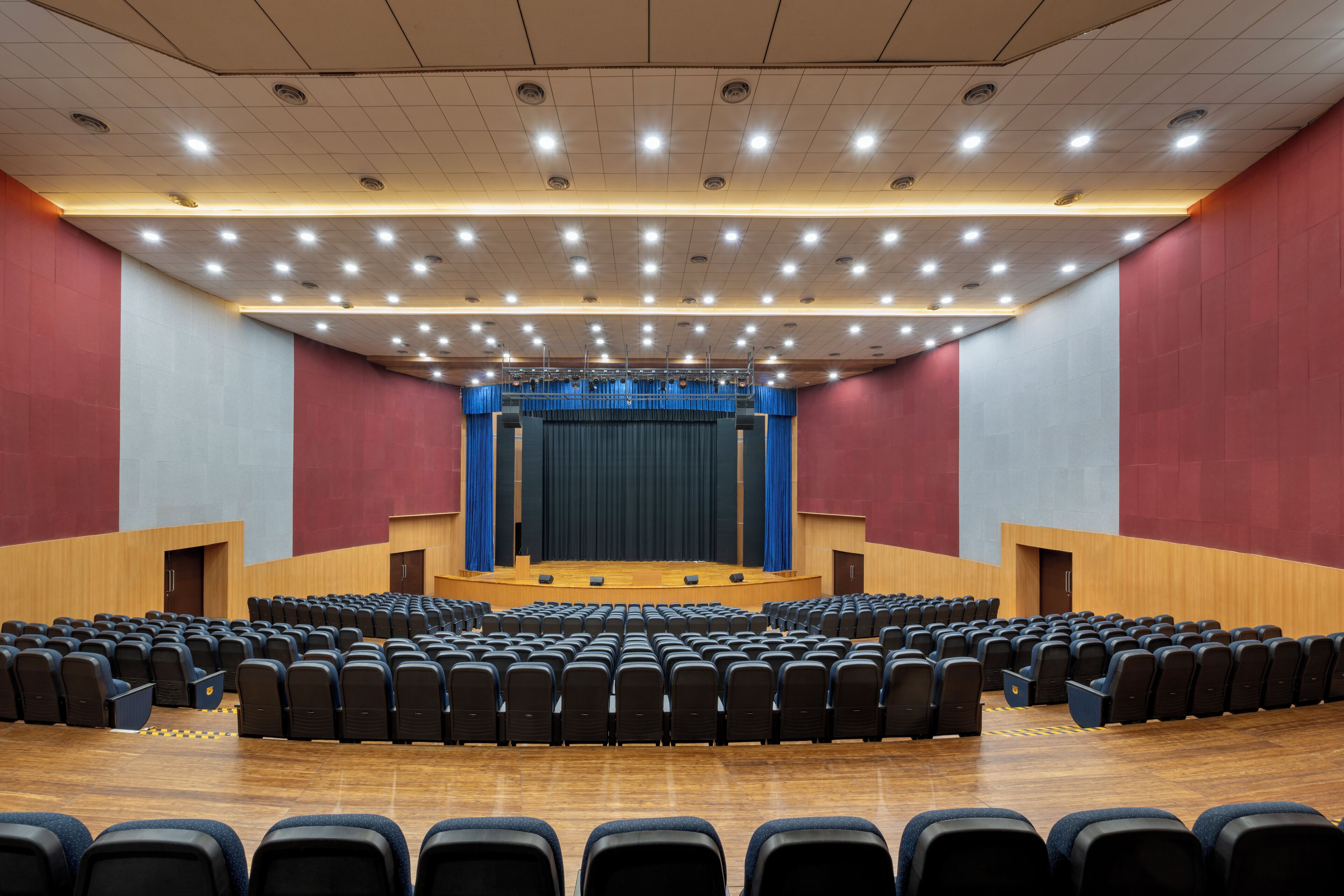 A large auditorium with rows of black chairs, a stage with a blue curtain, and a wooden floor.