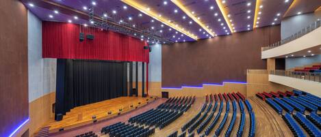 A modern auditorium with tiered seating, a stage with a red curtain, and a wooden floor.