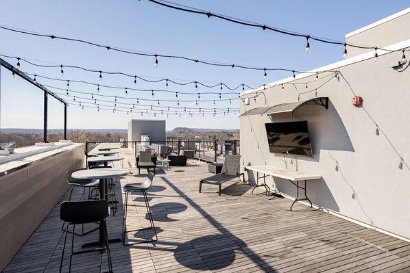 A rooftop terrace with tables, chairs, string lights, and a flat-screen TV.
