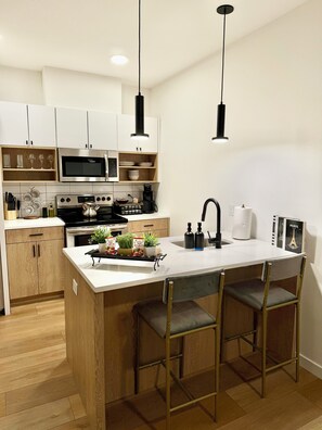 A modern kitchen with white cabinets, a central island with bar stools, a microwave, an oven, and a coffee machine.