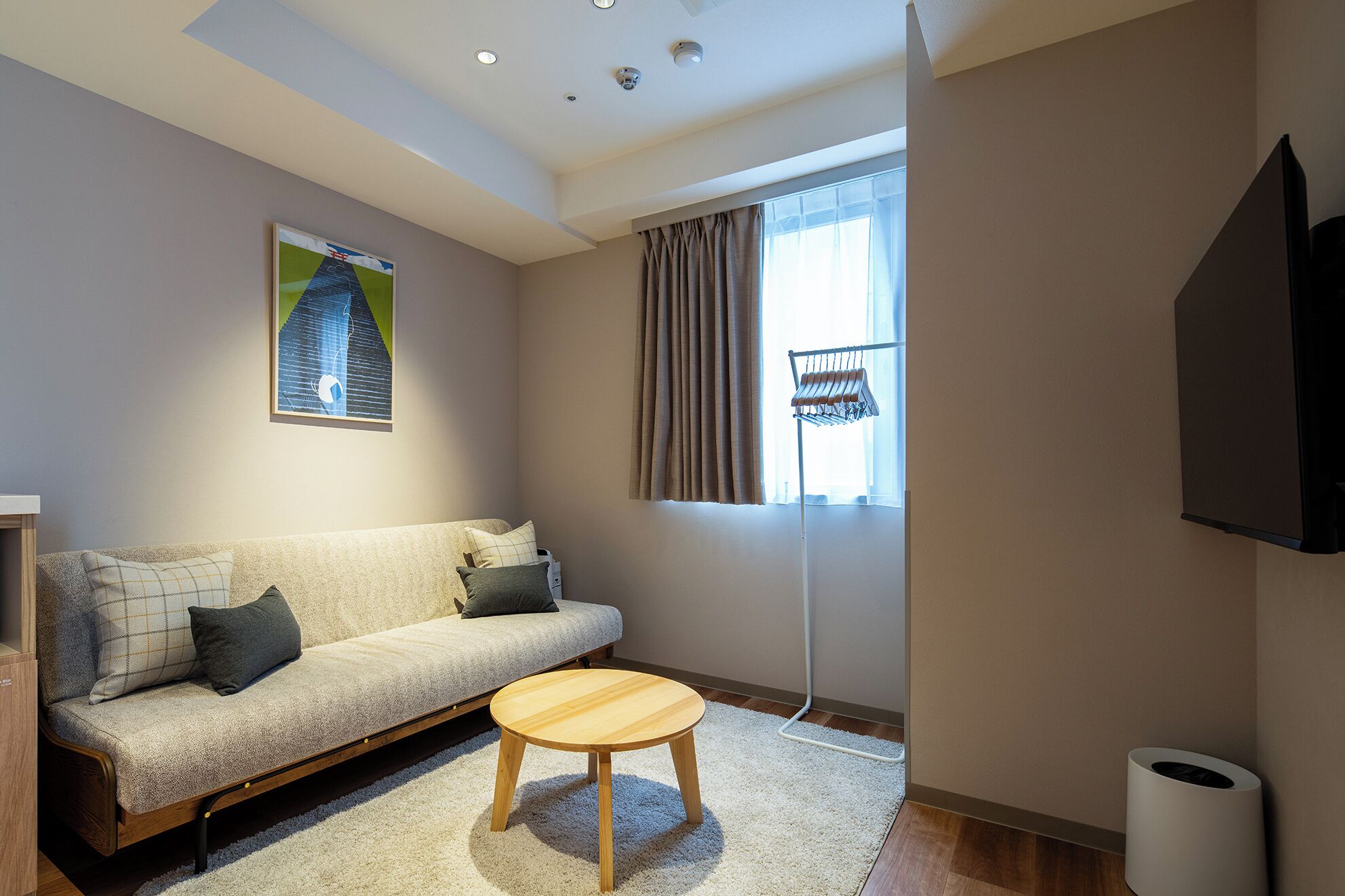 A modern living room with a sofa, a wooden coffee table, a painting, a window with curtains, and a mounted television.