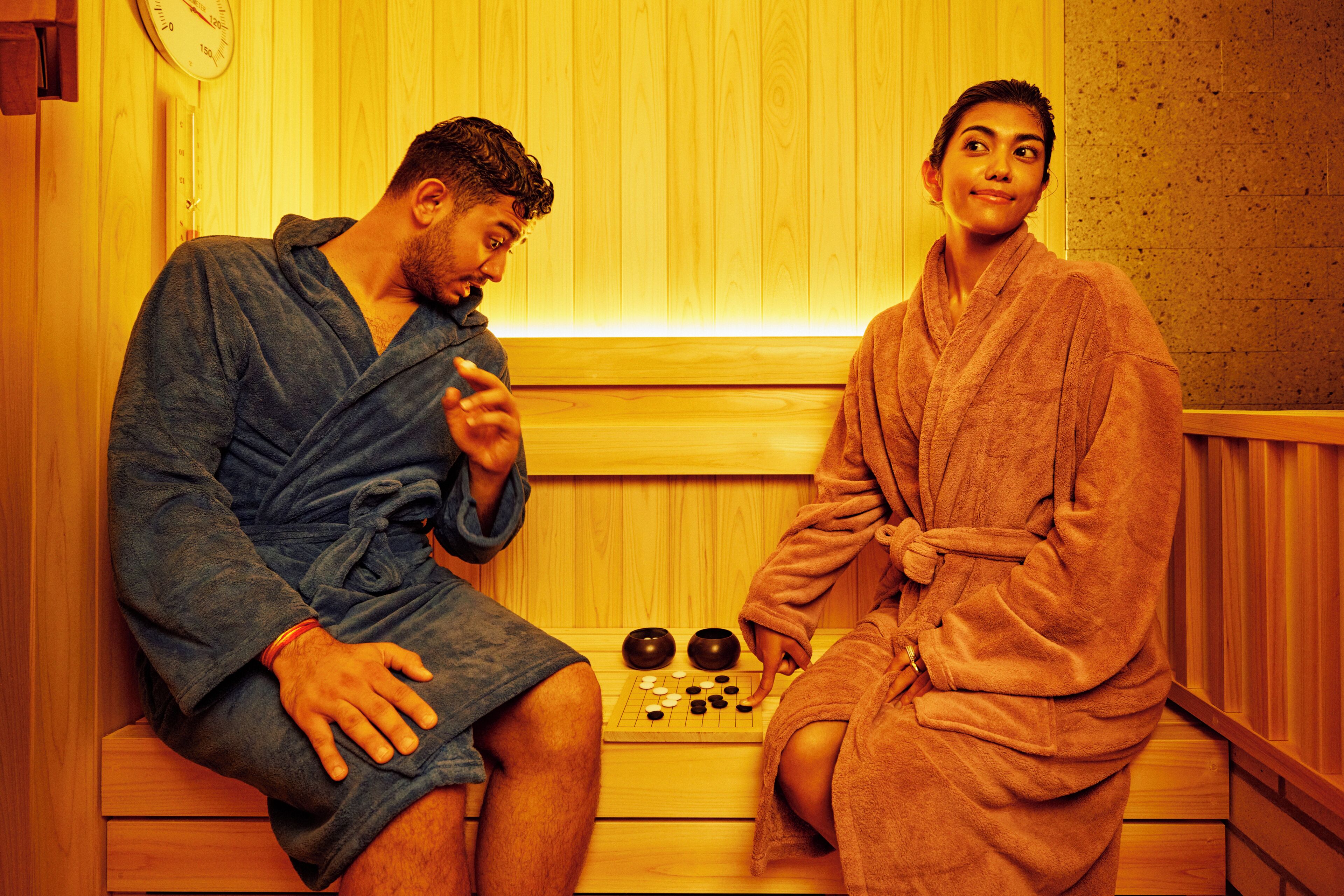 Two people in a sauna, one sitting and one standing, with a wooden bench and a small table with objects in front of them.