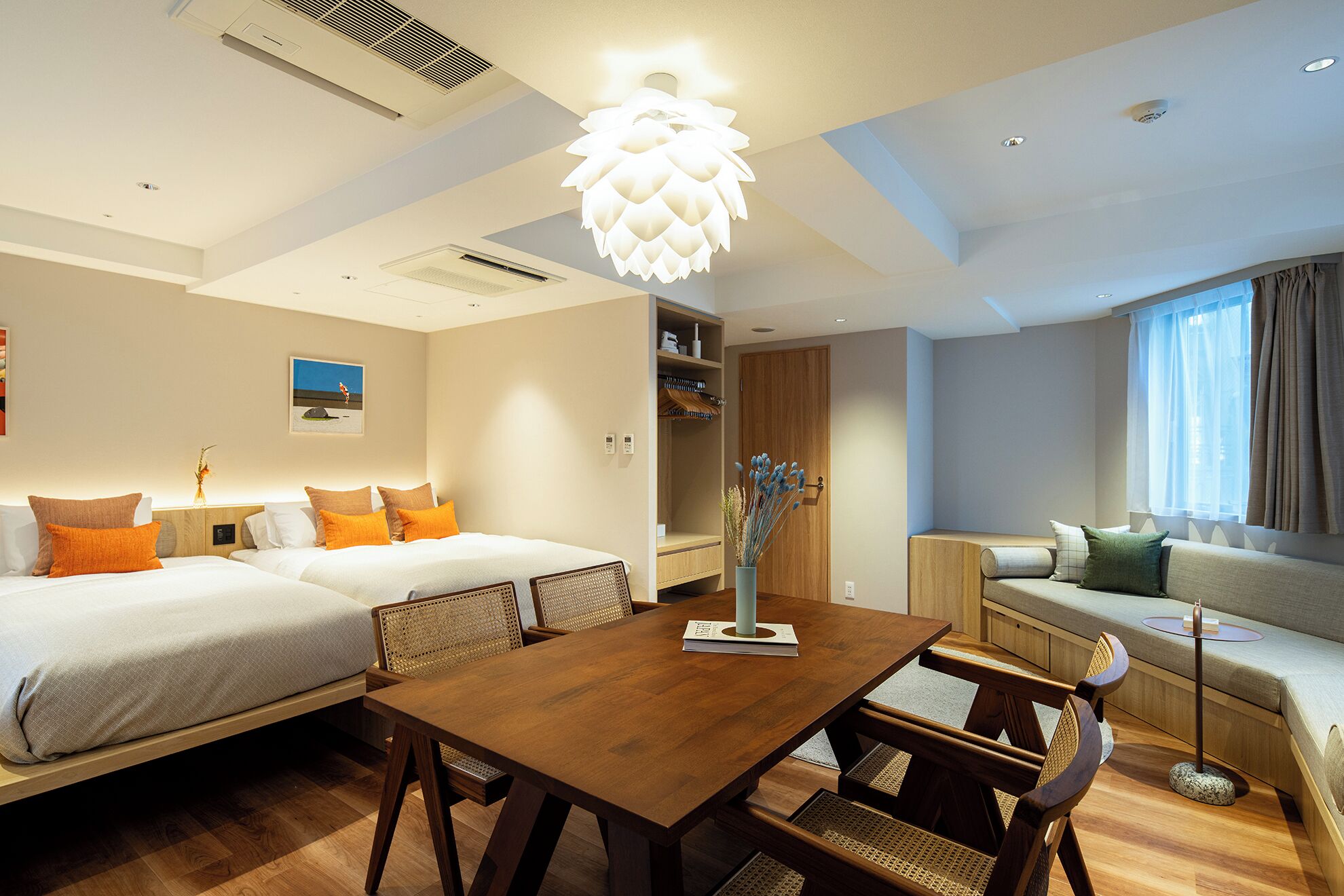 Ein modernes Hotelzimmer mit einem Holztisch, zwei Betten mit orangefarbenen Kissen, einer Couch und einem großen Fenster mit Vorhängen.