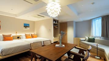Ein modernes Hotelzimmer mit einem Holztisch, zwei Betten mit orangefarbenen Kissen, einer Couch und einem großen Fenster mit Vorhängen.
