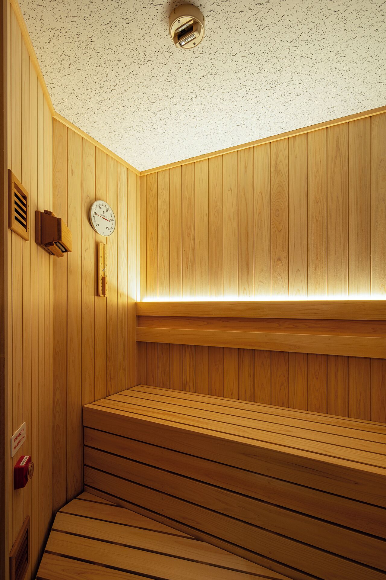 A wooden sauna with a sloped bench, a wall-mounted heater, a clock, and a ventilation vent.