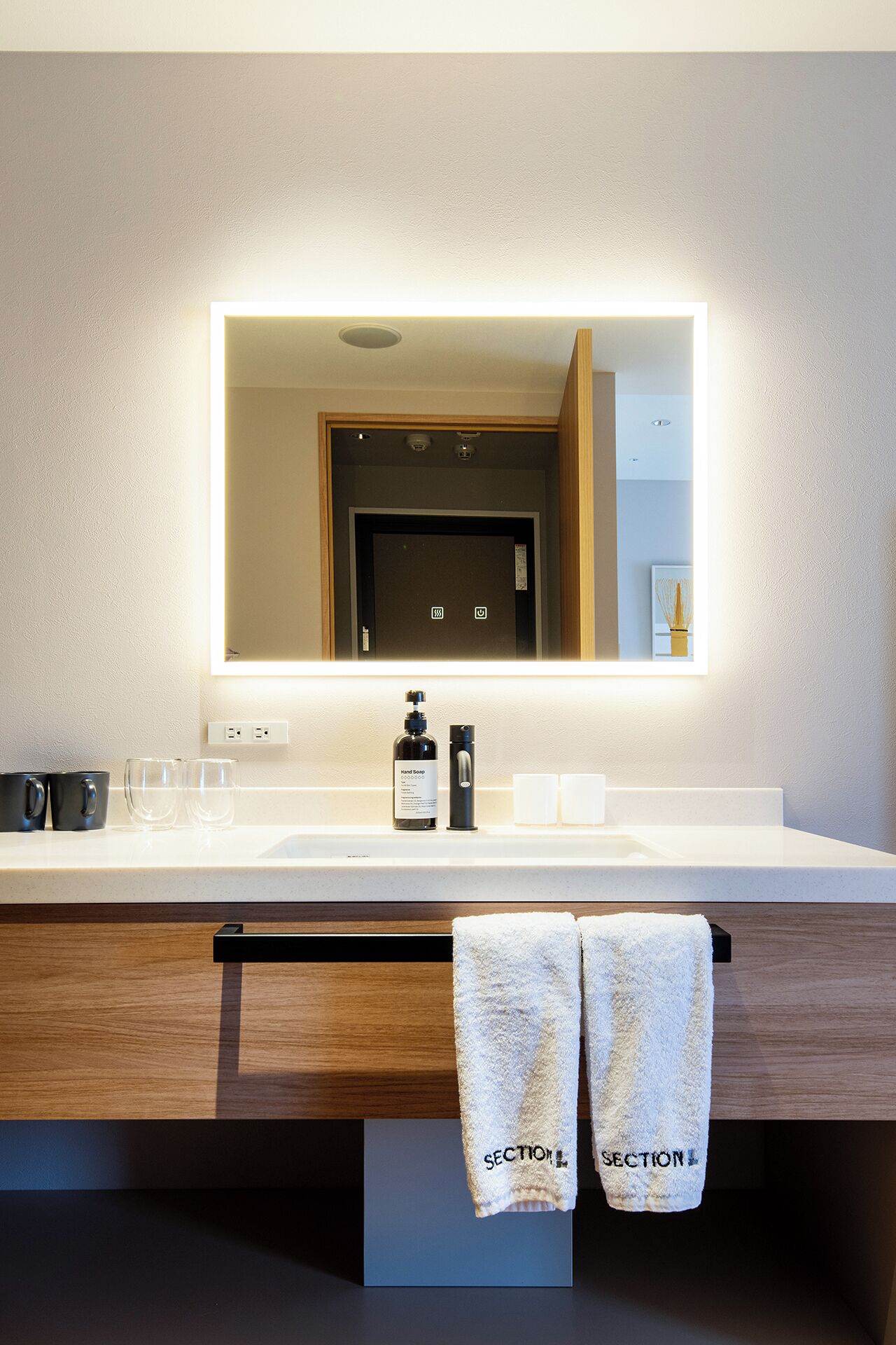 Un baño con un lavabo blanco, un espejo, un dispensador de jabón para manos y dos toallas colgadas en un perchero.
