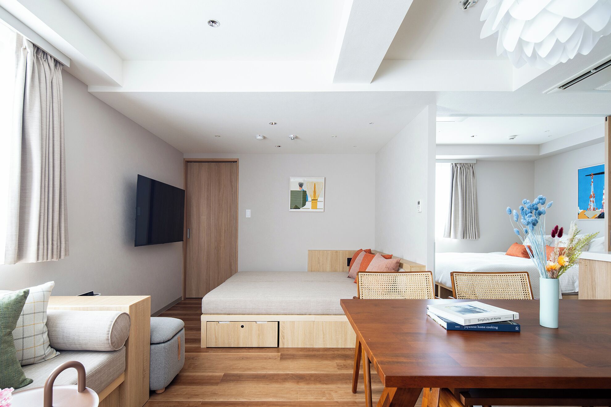 A modern, well-lit room with a wooden floor, a sofa, a bed, a dining table, and a TV mounted on the wall.
