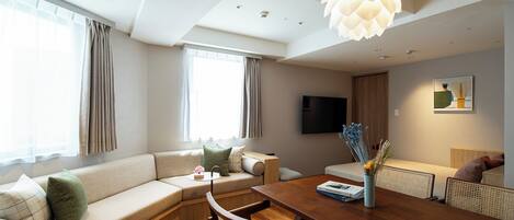 A modern living room with a sofa, dining table, and chairs. There is a flat-screen TV mounted on the wall and a decorative light fixture above the table.