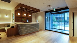 A modern reception area with a wooden reception desk, a seating area with a sofa, and large windows offering a view of the outdoors. - Section L Shimbashi West (Tokyo)