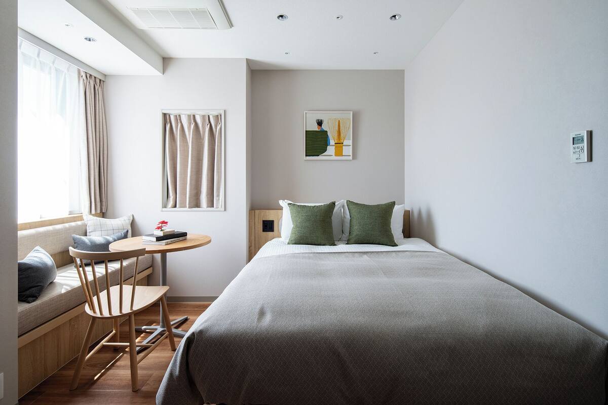 Quarto de hotel moderno com uma cama, uma mesinha com livros, uma cadeira e uma janela com cortinas.