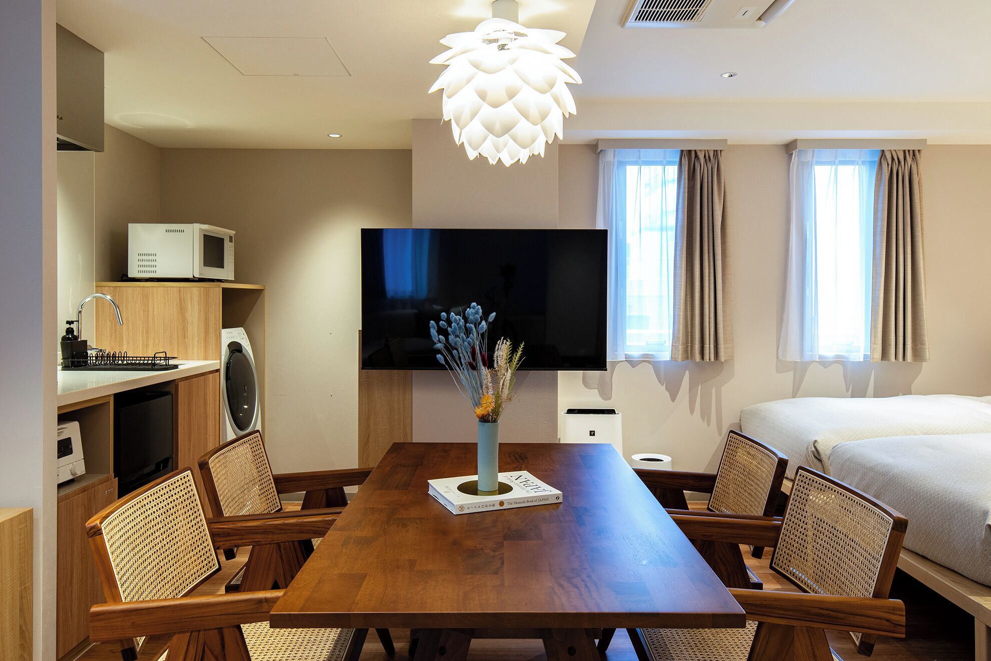 A hotel room with a wooden dining table, chairs, a flat-screen TV, a bed, a window with curtains, and a ceiling light fixture.