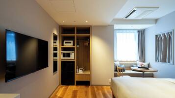 A hotel room with a flat-screen TV, a wooden wardrobe with built-in microwaves, a sofa, and a window with curtains.