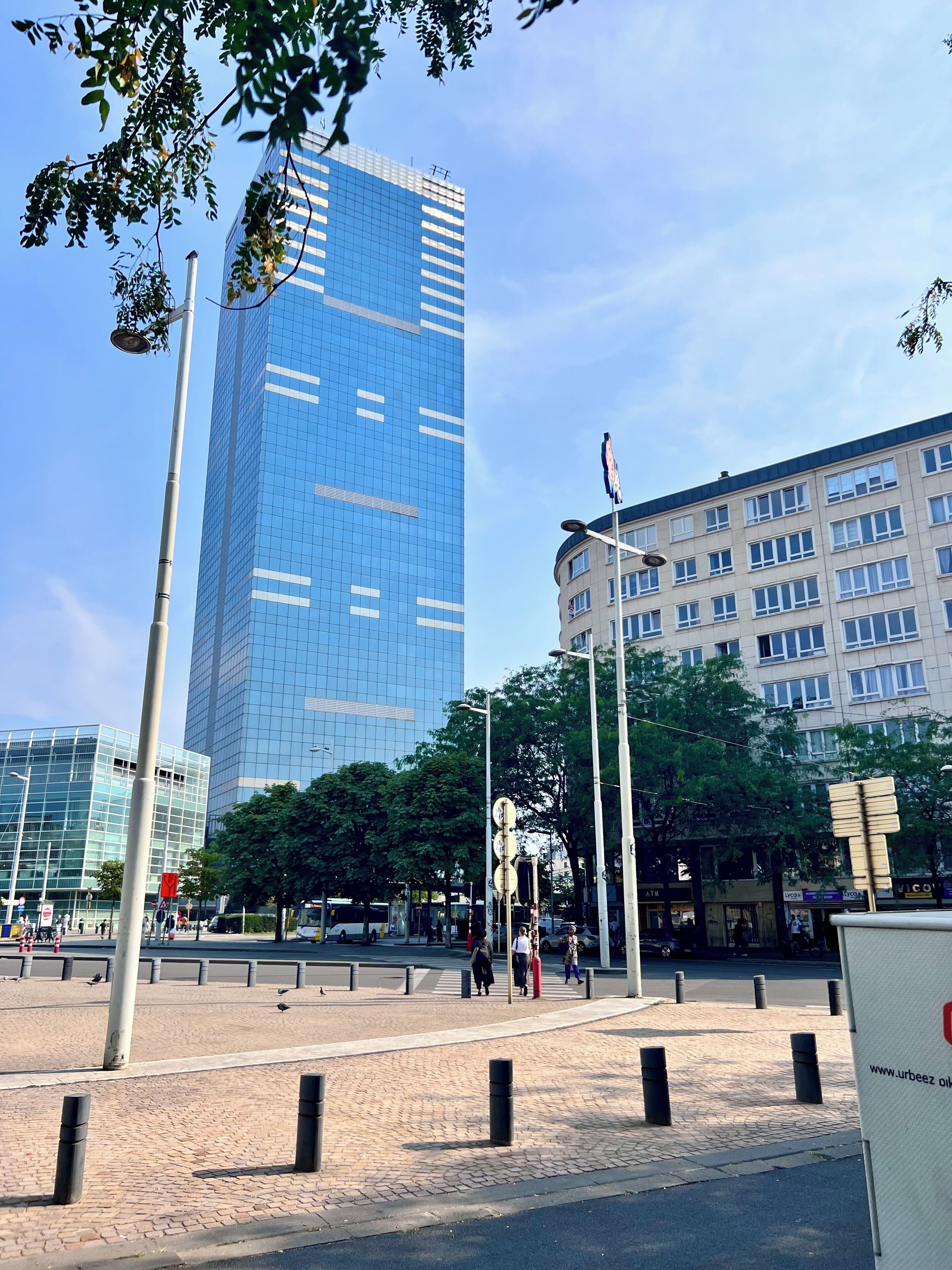 Une place urbaine moderne, avec un grand immeuble aux façades vitrées, un bâtiment circulaire doté de nombreuses fenêtres, et une rue bordée de bornes et fréquentée par des piétons.