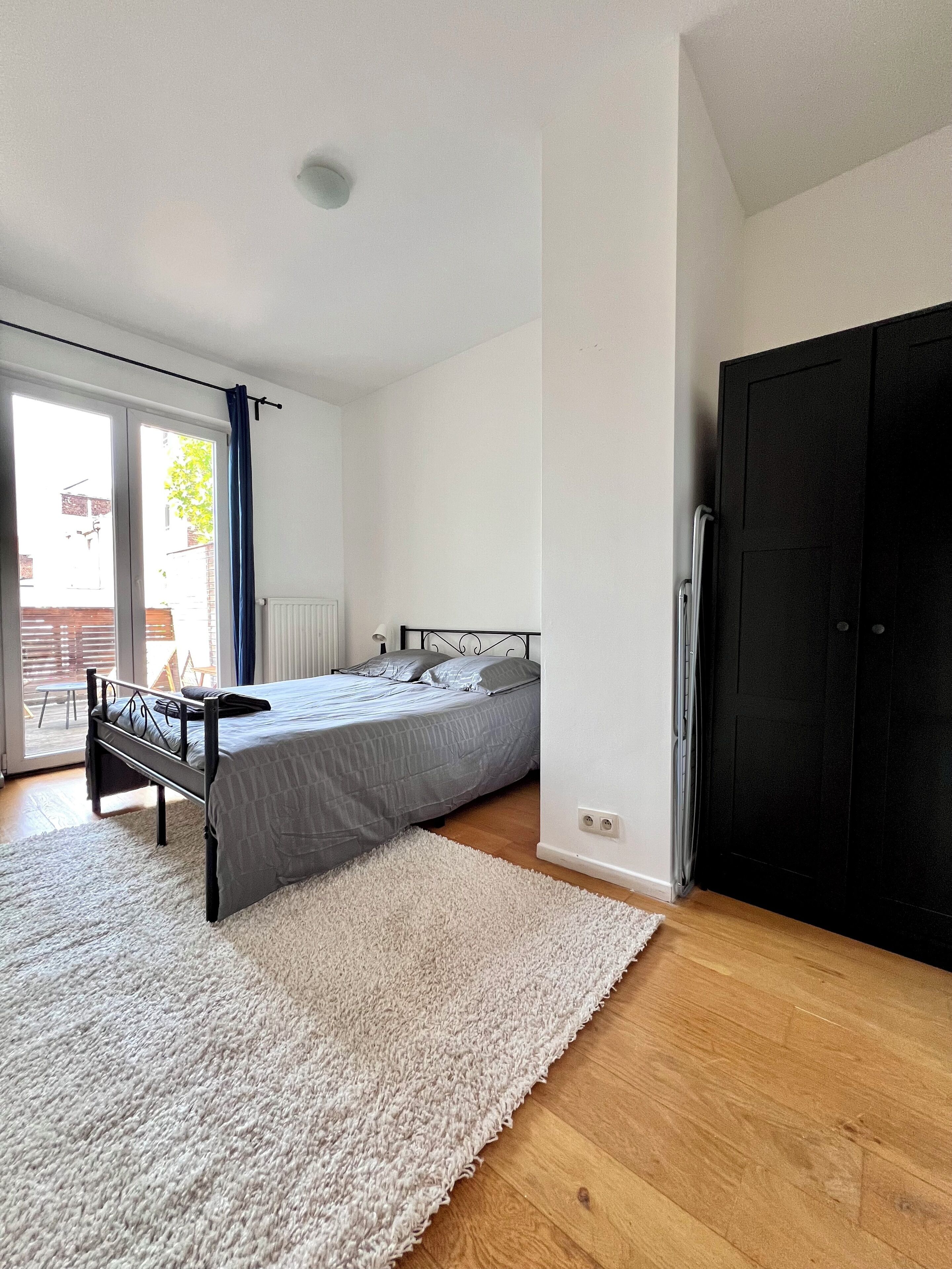 Un dormitorio con cama, alfombra blanca, suelo de madera, armario negro y puerta corredera de cristal al exterior.