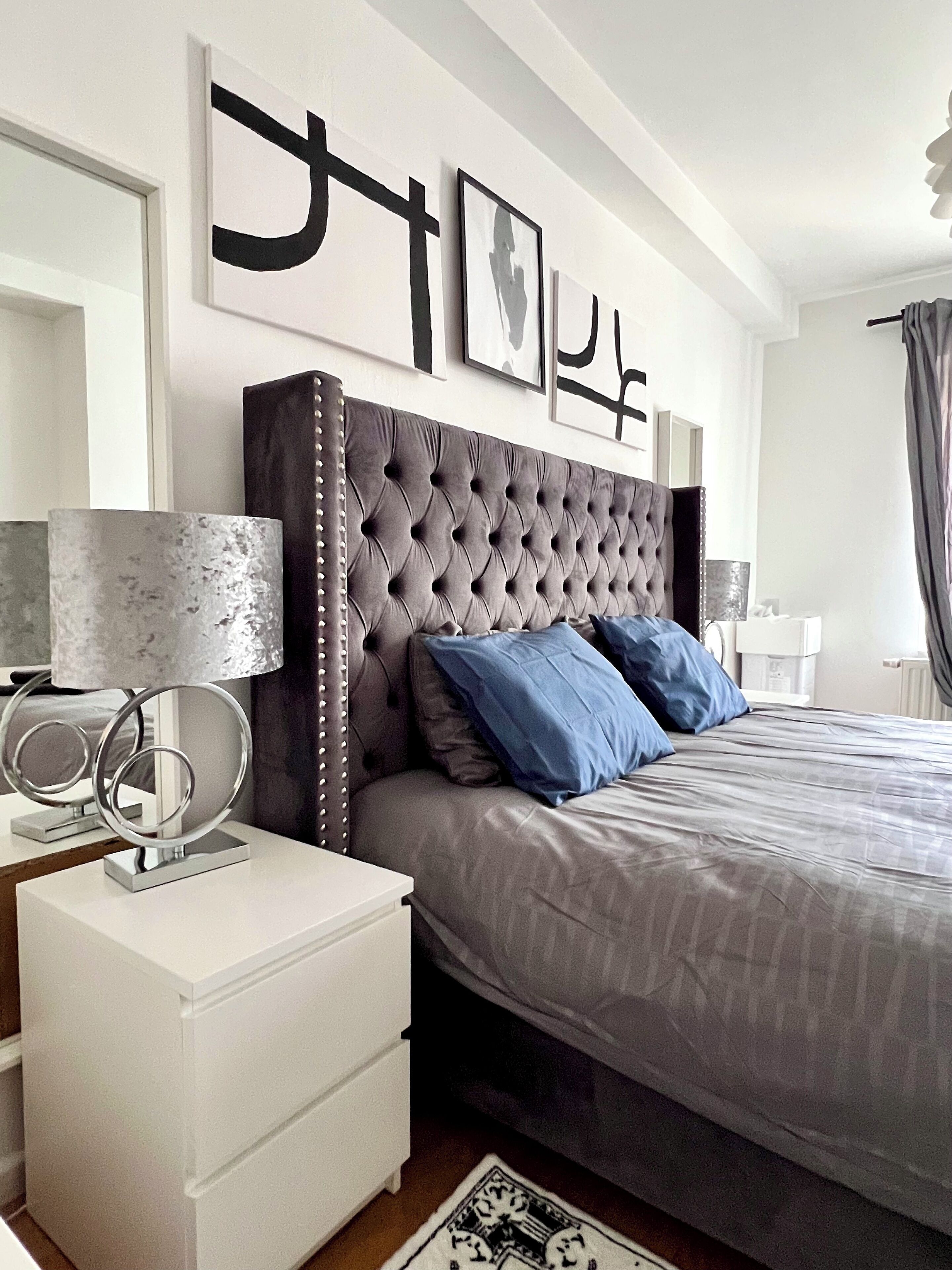 Une chambre à coucher moderne avec une tête de lit capitonnée, une couvre-lit grise et une table de chevet blanche avec une lampe.